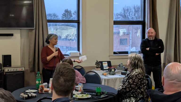 A woman announcing a website launch to a crowd of people in a reception room