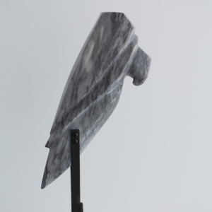 Sculpture en marbre gris en forme de tête de rapace, montée sur un support métallique noir surélevé, posé sur un socle en pierre blanche.