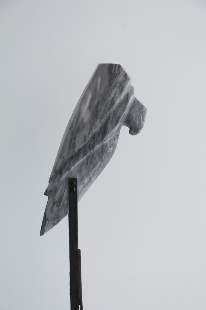 Sculpture en marbre gris en forme de tête de rapace, montée sur un support métallique noir surélevé, posé sur un socle en pierre blanche.