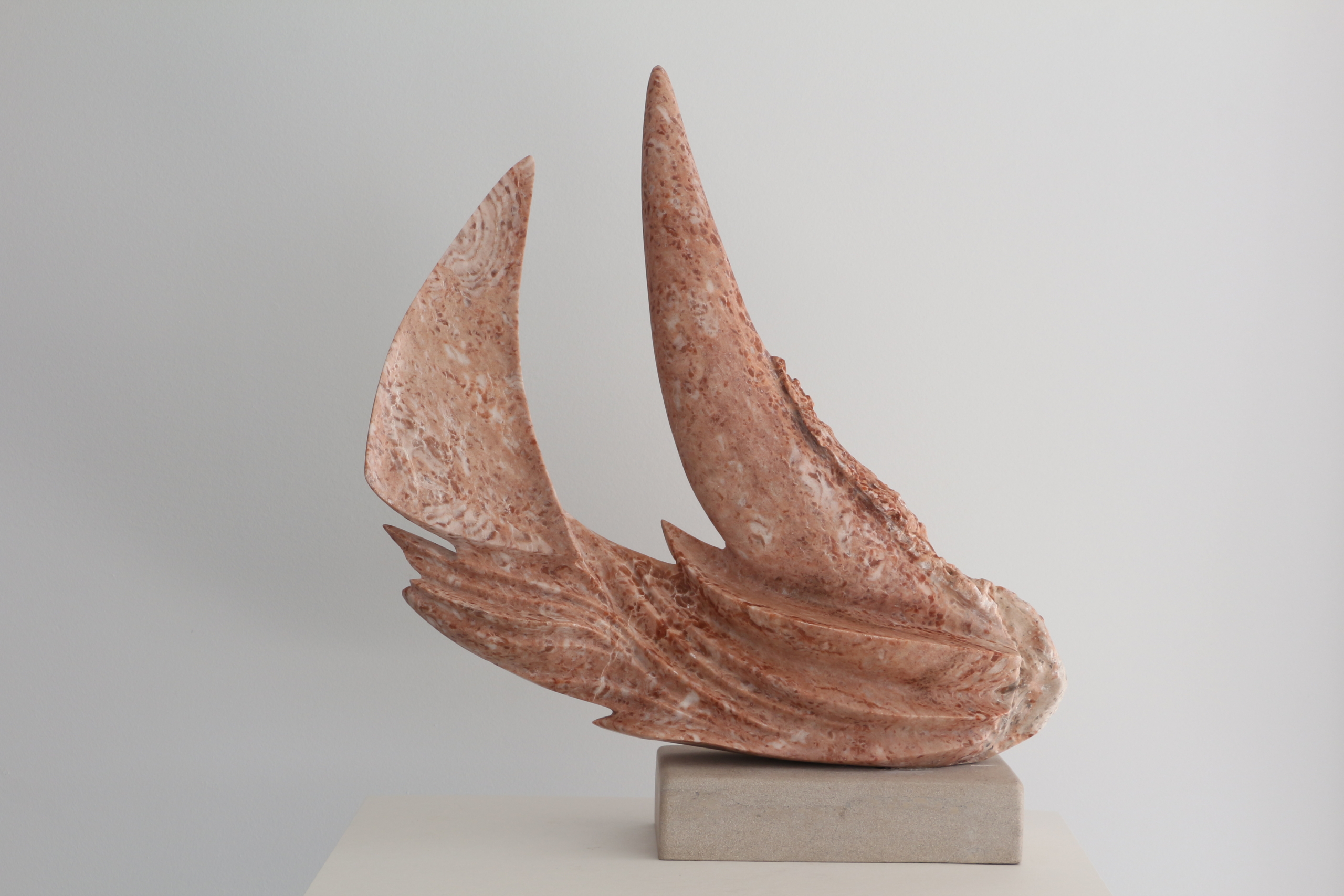 Sculpture abstraite en pierre rose veinée de terre cuite, en forme de voiles ou d'ailes courbées, posée sur une base en pierre beige.